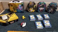Photo showing four metal Pokemon collectible tins featuring Pikachu, Mew, Jolteon, Zapdos, Lugia and Articuno. About 250 Pokemon cards in stacks with blue Pokeball card backs. Small plush Pokeball keychain and a yellow Pikachu figure with visible wear.