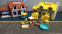 Photo showing two vintage Fisher Price play houses: yellow 1969 house and brown 1980 house, with multiple small plastic furniture pieces and figures arranged in front