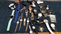 Image showing approximately 30 watches of various styles and sizes laid out on a dark fabric background, including metal, leather, and fabric straps in various colors; some watches clearly vintage and used.