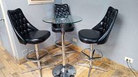 Three black vinyl diamond-tufted bar stools surrounding a round clear glass top chrome pedestal bar table on wooden floor.