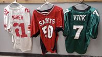 Front view of three vintage NFL jerseys hanging on hangers, showing New York Giants Tiki Barber white #21, New Orleans Saints red #50, and Philadelphia Eagles Michael Vick green #7.