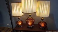 Front view of the three pumpkin-shaped amber blown glass lamps with pleated cream shades on a wooden table, lamp cords visible.