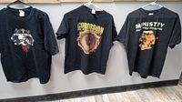 Three black concert T-shirts hung on hangers displaying front graphics for Necrotek, Corrosion of Conformity, and Ministry bands.