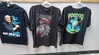 Three black cotton concert t-shirts hanging on a wall: Phil Collins, Metallica, and Voodoo Fest designs visible.