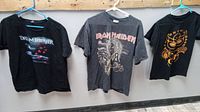 Front view of three t-shirts hanging on a rack showing Dream Theater, Iron Maiden, and Zac Brown Band concert t-shirts