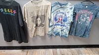 Four rock band T-shirts hanging on hooks against a gray wall, showing their front graphics.