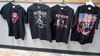 Photo of all four black large rock band T-shirts hanging on hangers, featuring Rolling Stones, Joe Cocker, John Kay Steppenwolf, and KISS with makeup.
