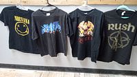 Four black rock band T-shirts hanging on hangers, front facing. From left to right: Nirvana, Black Sabbath, Queen, Rush.