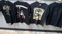 Photo showing four vintage black rock T-shirts featuring AC/DC, Jimi Hendrix, Black Sabbath, and Rolling Stones on hangers.