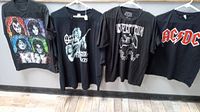Four black rock band t-shirts hanging on hangers on a wall; KISS, Ozzy Osbourne, Led Zeppelin, and AC/DC visible.