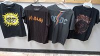 Four black rock band T-shirts hanging on hangers showcasing front designs for Sublime, Def Leppard, AC/DC, and Ramones.