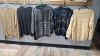 All five sweaters hanging on hangers against a wooden backdrop on a rail: cream cable knit, charcoal solid, two patterned sweaters in gray and blue, and another cream sweater.