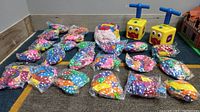 Overview of 23 sealed bags of colorful polka-dotted balloons spread on carpeted floor with 2 yellow balloon blower pumps with blue handles and face designs.