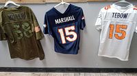 Three Denver Broncos jerseys hung on hangers. From left to right: green Von Miller number 58 throwback, navy blue Brandon Marshall number 15 women's large, white Tim Tebow number 15 women's medium.