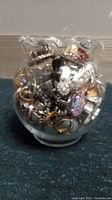 Clear glass jar filled with a variety of multicolored and metallic costume rings, showing diverse styles including enamel, faux stones, and plating.