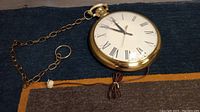 Large vintage pocket watch style wall clock displayed from an angle showing attached chain and power cord.