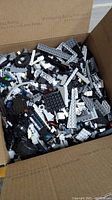 Open USPS Priority Mail box filled with assorted black, white, and gray LEGO bricks and parts, showing many different pieces including plates and wheels.