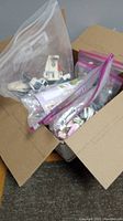 Open cardboard box revealing multiple clear plastic bags filled with various LEGO pieces and small builds, some bags sealed with purple zip lock.