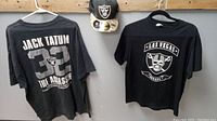 Lot displayed showing the Jack Tatum 32 The Assassin black t-shirt, Las Vegas Raiders skull logo t-shirt, and Raiders on field cap hung on wall.