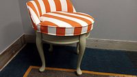 Vanity swivel chair with cream wooden cabriole legs and orange and white striped upholstery on a carpeted floor near wall.