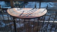 Side angle of a semi-circular mosaic tile portable table mounted on an outdoor metal railing overlooking a snowy landscape.