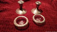 Two sterling silver candle holders and two matching sterling silver ashtrays with floral embossed rims set against a red fabric background.
