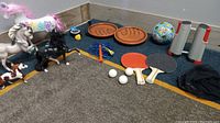 Photo showing six horse figurines, horseshoe-shaped paddles, ping pong paddles, balls, portable net, jump rope, and basketball all on carpeted floor corner.