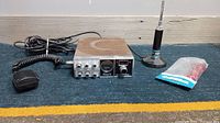 Photo showing vintage CB radio unit, microphone to left, magnetic antenna rod on right, and small packaged plug on floor