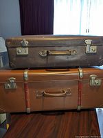 Side view of two vintage suitcases stacked, showing dark brown suitcase on top with metal clasps and twisted handle, and tan suitcase below with leather straps.