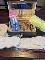 Full shot of the vintage style black make-up box open showing interior with mirror, Chanel perfume bottle inside, a nail file kit, a yellow shower scrubber, and two white slippers in packaging in front.