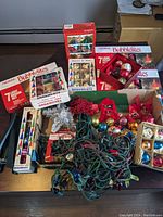 Photo of multiple boxed NOMA Bubblelites light sets and several boxes of assorted Christmas ornaments including glass balls, grape clusters, and decorative holly berry sprays on a table.