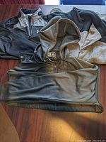 Black Under Armour hooded shirt jacket and heather grey hoodie laid flat on table alongside fleece vest hung on a hanger