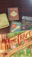Photo showing four Minecraft handbooks with pixel pickaxe designs and colorful Minecraft poster laid on a surface.
