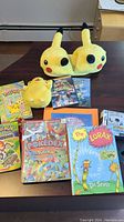 Overall view of lot items including Pikachu slippers, Pikachu plush, Pokemon handbooks, DVDs, and other books arranged on a dark wooden table.