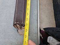 Close up of box newel post end showing paint chipping, held with measuring tape approx 15 cm wide.