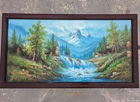 Full frontal view of the framed painting showing the mountain, waterfall, trees, and cabin landscape.