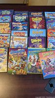 Top-down view of 19 Scooby Doo DVD covers laid out on a dark table, showing a colorful array of cases with various titles and artwork.