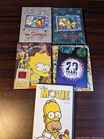 Five DVDs from The Simpsons series arranged on a wooden surface. Covers show colorful cartoon artwork representing different seasons and the movie.