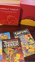 Simpsons World book with red cover shows stylized line art of Homer, Lisa, and Maggie Simpson. Three comic books visible beneath including The Simpsons Treehouse of Horror and Simpsons Comics with family on cover.