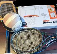 Photo showing the volleyball, mini basketball hoop box, and several rackets arranged on a dark surface.