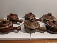 Front view of 6 brown ceramic pots with lids and side handles arranged on wooden surface.