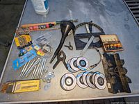 Woodworking tools and accessories on a metal surface including clamps, grinding wheels, various hardware, a plaster mixer attachment, black funnel, and metal measuring tools.