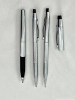 Three silver tone Cross pens and pencils displayed vertically, showing capped ballpoint pen with black grip, mechanical pencil, and capped pen or pencil.
