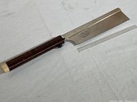 Side view of the Takuma craft saw showing the full blade and wood handle wrapped in molded dark brown plastic. A clear ruler lies next to the blade showing approximate length.