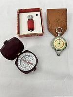 Three vintage items including a brown leather-cased altimeter/compass, a maroon folding compass, and a boxed Audubon bird call with red wooden handle.