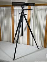 Full view of Swift Mark II Zoomscope mounted on Leitz Tiltall tripod standing fully extended on a white surface with a backdrop of vertically pleated curtains.