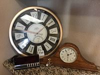 Two analog clocks displayed on a granite shelf: One large round analog clock with a metallic frame and mixed numerals; One wooden mantel La Crosse clock with white face and Arabic numerals