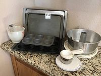 Photo showing glass measuring cup, two muffin tins (one large and one mini), two baking sheets, aluminum tube cake pan with plastic base, and measuring cups on a ceramic coaster.