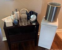 Full set of bathroom accessories shown on dark wooden stand and floor, includes glass perfume bottle, soap dispensers, hand mirrors, tissue box, acrylic organizer, double cups, small waste bin, and clothing basket.