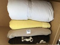 Three folded blankets stacked: white down comforter, yellow fleece blanket, beige fleece blanket, dark brown patterned fleece blanket at bottom.